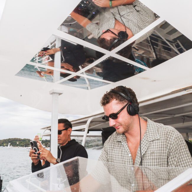 Two men on a boat, both wearing sunglasses and headphones. One man stands at a DJ deck, seemingly mixing music, while the other operates a camera. The scene is set on water with boats and land in the background.