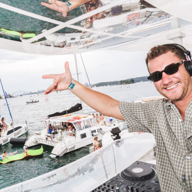 A smiling DJ wearing sunglasses and headphones performs on a boat. Behind him, people relax on yachts and inflatables in the water under a partly cloudy sky, creating a lively party atmosphere.