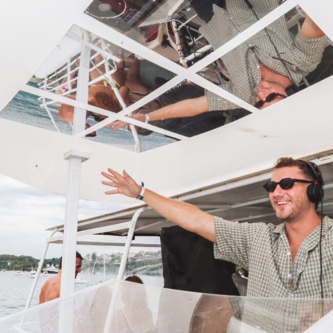 A smiling man in sunglasses and headphones spreads his arms wide while DJing on a boat, with his reflection visible in mirrored panels above. Boats and water are in the background.
