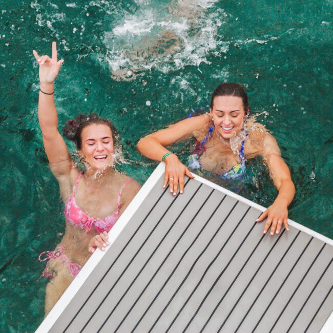 Two smiling women in bikinis enjoy swimming in clear green water, holding onto the edge of a floating dock. One woman raises her hand, making a "rock on" gesture.