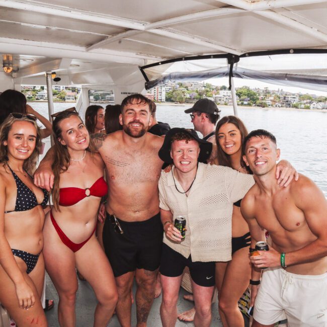 A group of young adults in swimwear smile and pose together on a boat, with drinks in hand. The boat is on a body of water with buildings and greenery visible in the background.