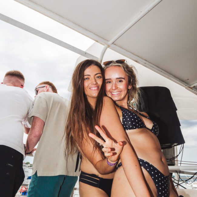 Two young women in black polka-dot bikinis smile and pose, one flashing a peace sign, on a boat with other people and a busy waterfront in the background under a partly cloudy sky.