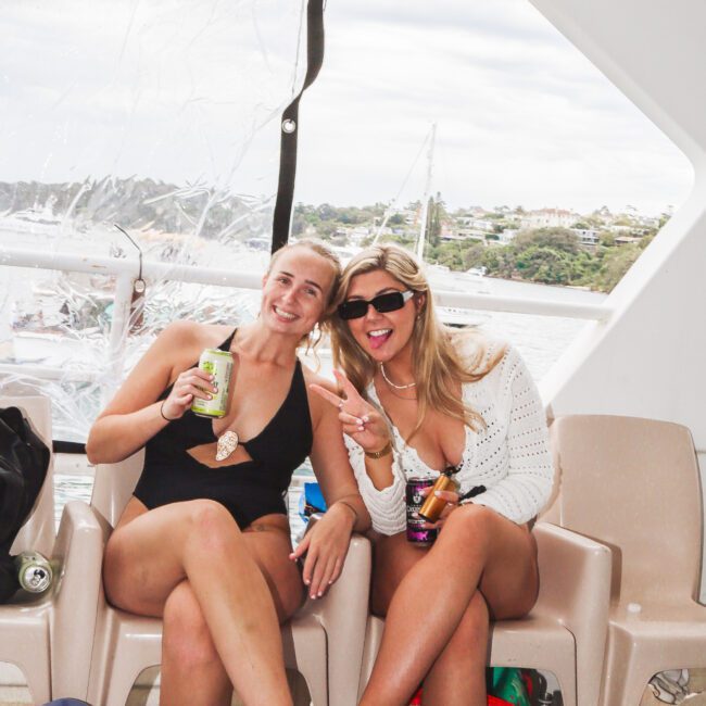 Two women in swimsuits sit on chairs on a boat, smiling and holding drinks. The background shows water, boats, and greenery. One woman makes a peace sign and they both appear to be enjoying a relaxed day.