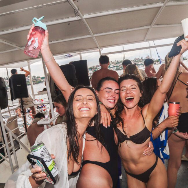 Three women in swimsuits smile and pose with drinks, celebrating on a crowded boat. Other people in swimwear are in the background, and the boat is on the water near a city or shoreline. The mood is festive and joyful.