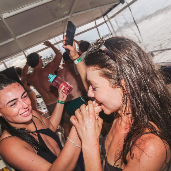 Two women in swimsuits smile and dance closely on a boat party, surrounded by other people enjoying themselves. One woman holds a phone while another has wet hair, suggesting they have been swimming. The ocean is visible in the background.
