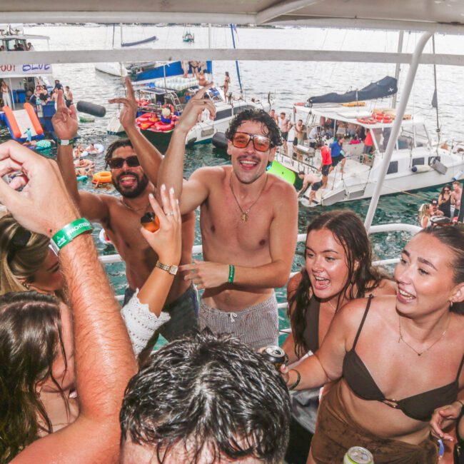 A group of young adults dance, smile, and raise drinks while partying on a boat. Other boats and people are visible in the background, with the ocean and a festive atmosphere all around.