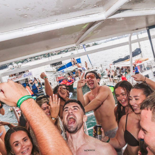 A group of people in swimsuits dance and celebrate on a crowded boat, raising their arms and smiling, with other boats and water visible in the background.