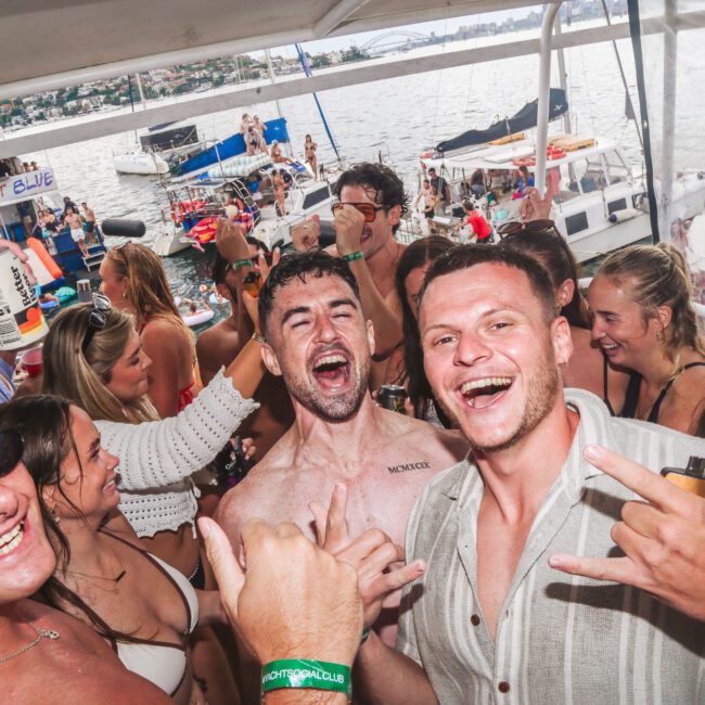 A group of young adults in swimsuits laugh, cheer, and raise drinks on a crowded boat during a sunny day, with more boats and people visible on the water in the background.
