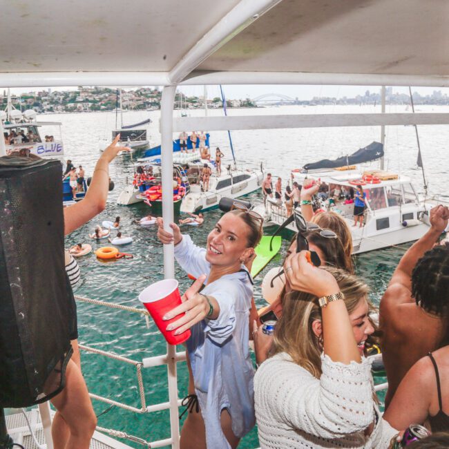 A group of people dance and smile on a boat, holding drinks, with several boats and people in the water in the background on a sunny day. The scene is festive and energetic.