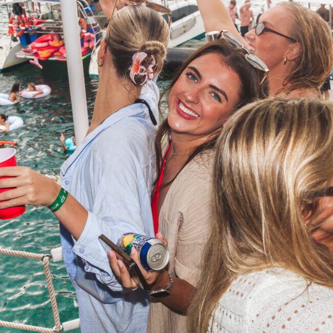A group of women on a boat party smile and dance, holding drinks. Other boats and people floating on colorful inflatables are visible in the water behind them. It’s a lively, outdoor summer scene.