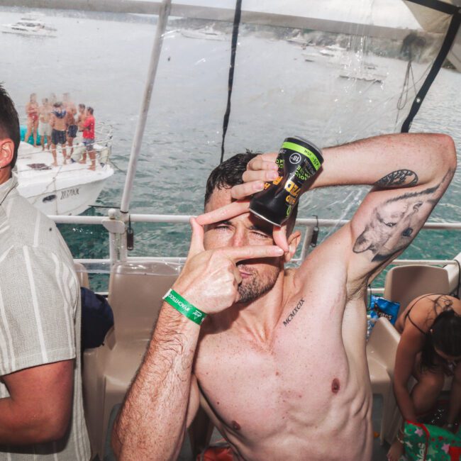 A man poses playfully on a boat, holding a drink and framing his face with his hands. Other people are nearby, one sitting and another looking at the camera. Boats and water are visible in the background.