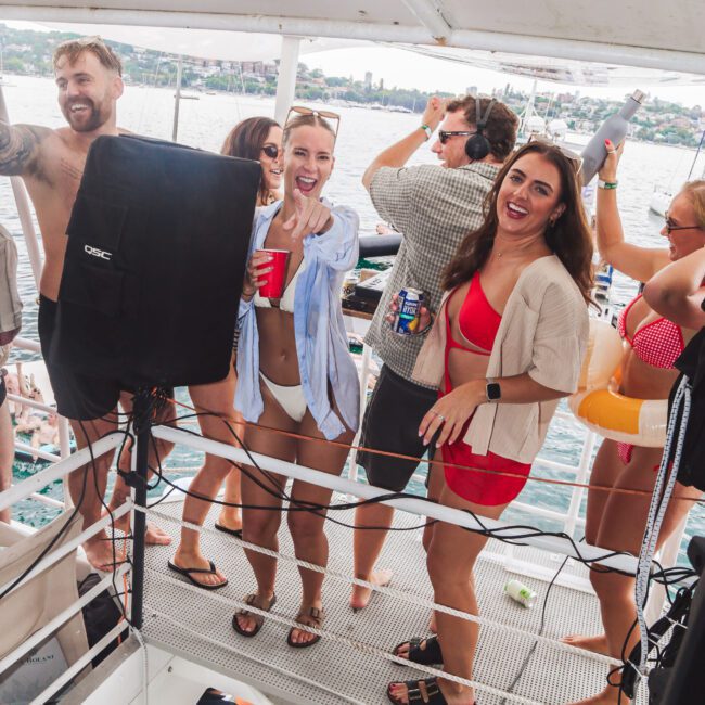 A group of people in swimsuits and casual summer clothes happily dancing and holding drinks on a boat, with water and a city skyline in the background. Some are smiling at the camera and enjoying the lively atmosphere.