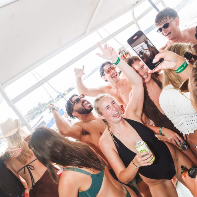 A group of young adults in swimsuits dance and smile on a boat, some holding drinks and raising their hands. One person takes a selfie while others cheer and laugh, creating a lively, festive atmosphere.