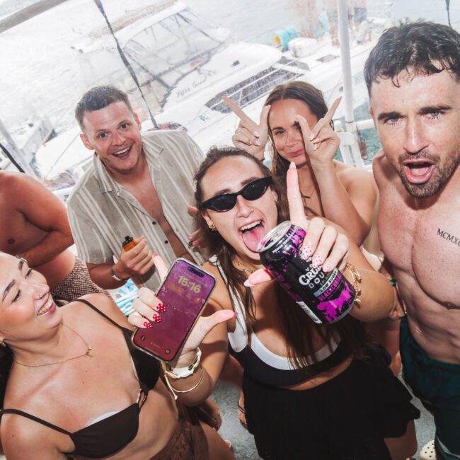 A group of young adults in swimwear enjoy a lively boat party, smiling, holding drinks, and posing for the camera with the ocean and other boats visible in the background.