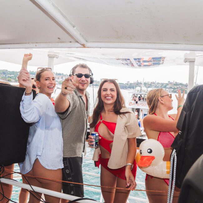 A group of people on a boat, smiling and dancing. One person points towards the camera, another wears a red swimsuit and holds an inflatable duck. The background shows water and other boats.