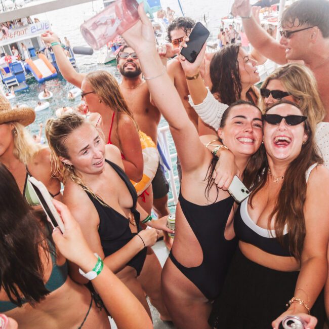 A group of young adults in swimwear smile, dance, and raise drinks while crowded together at a lively boat party on the water. The atmosphere is festive and energetic under sunny skies.