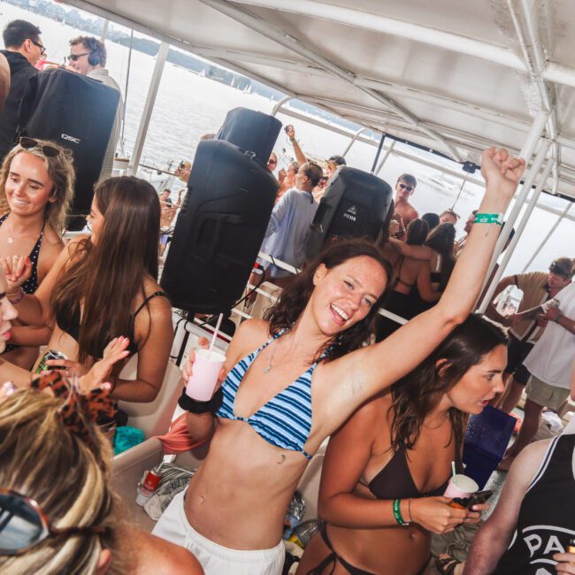 A group of people in swimwear enjoy a lively boat party, with a woman in the center raising her arm and smiling, while others chat, hold drinks, and dance under a covered deck with water in the background.
