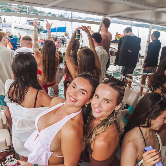 A group of young adults in swimwear enjoy a lively party on a boat, smiling, dancing, and holding drinks, with a scenic waterfront and other boats visible in the background.