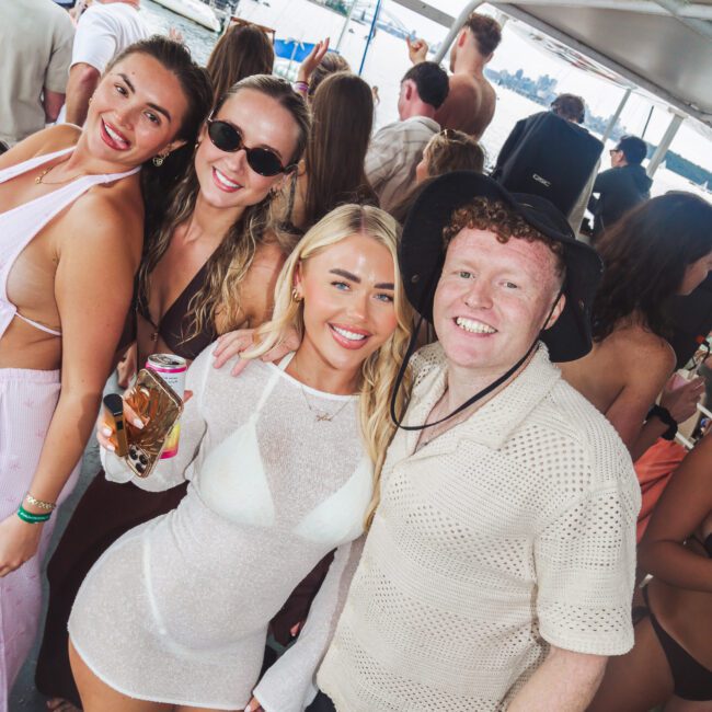 Four people smile and pose together at a crowded boat party, with water and other boats visible in the background. They wear summer clothes, sunglasses, and swimwear, and one holds a drink.
