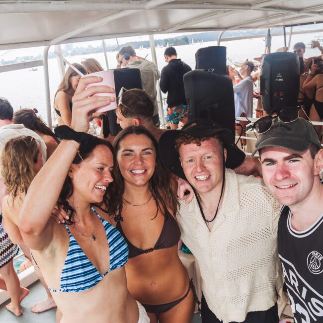 Four smiling people in swimwear pose for a selfie on a crowded boat, with others mingling in the background. The atmosphere is lively, and the boat is on the water with more boats visible nearby.