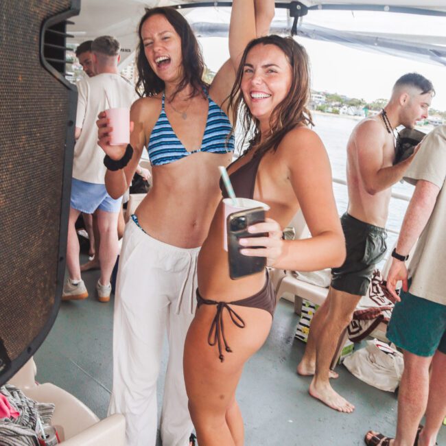 Two women in swimwear laugh and raise their joined hands while holding drinks on a boat, surrounded by other people relaxing and enjoying a sunny day near the water.