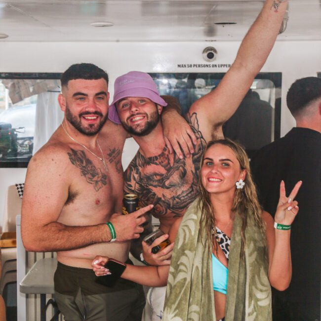 Three people smile and pose for a photo at a party. Two shirtless men with tattoos stand close together; one wears a purple bucket hat. A woman in front flashes a peace sign and holds a drink, wrapped in a towel.