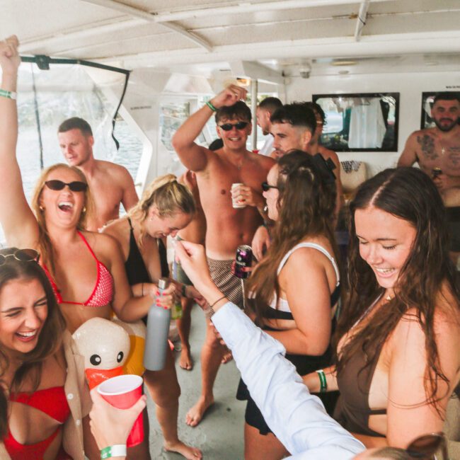 A group of young adults in swimsuits dance, laugh, and hold drinks while enjoying a lively party on a boat. Some people raise their arms in excitement, and there is an inflatable duck visible among the crowd.