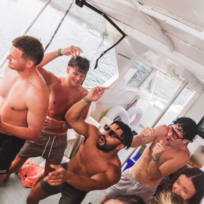 A group of young adults in swimsuits dance and laugh together on a boat. Some are wearing sunglasses, and one person is holding a phone to take a selfie. The atmosphere appears fun and lively.