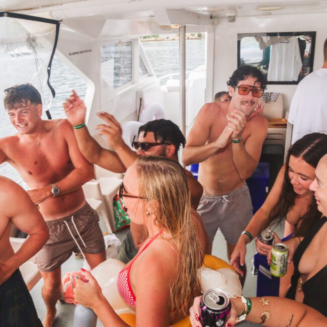 A group of young adults in swimwear dance, laugh, and hold drinks while enjoying a lively party on a boat. Sunlight streams in and the background shows water and part of the boat's interior.