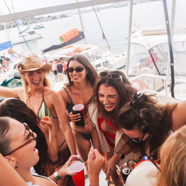 A group of young women in swimsuits laugh and cheer together on a boat, holding drinks, with other boats and people visible on the water in the background. It’s a lively, sunny day party scene.