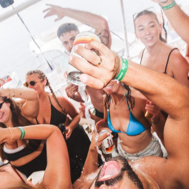 A group of people in swimwear are partying energetically on a boat, splashing drinks and cheering with cans in hand. The atmosphere is lively and festive, suggesting a summer celebration.