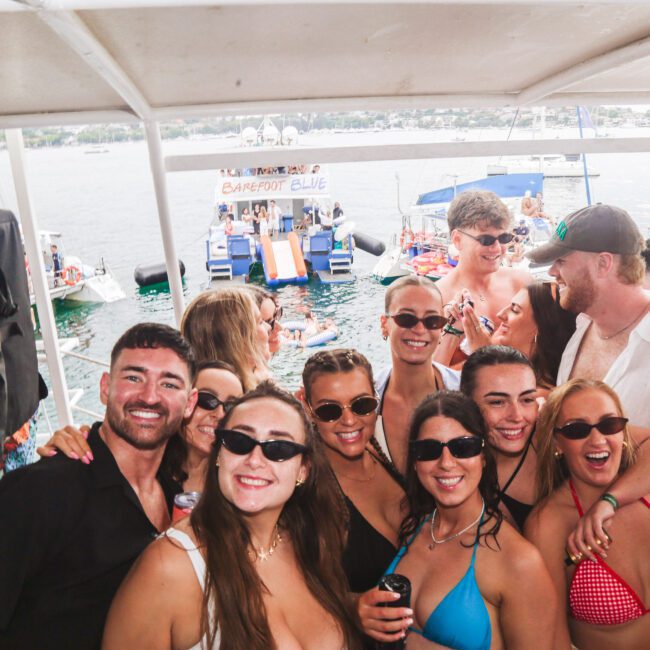 A group of young adults in swimsuits and sunglasses smile and pose for a photo on a boat, with other boats and people visible in the water and background. The scene is bright and festive, suggesting a party atmosphere.