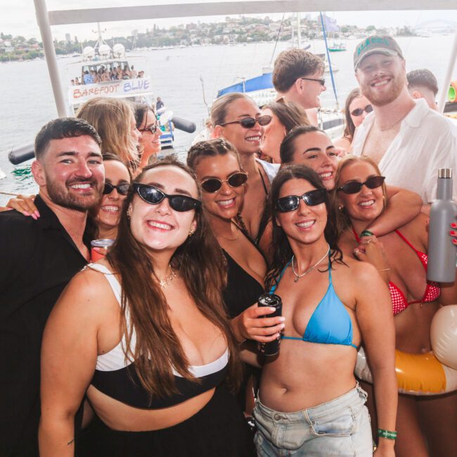 A group of smiling young adults in swimwear pose for a photo at a lively boat party. Some wear sunglasses, and one person holds an inflatable duck. Water and cityscape are visible in the background.