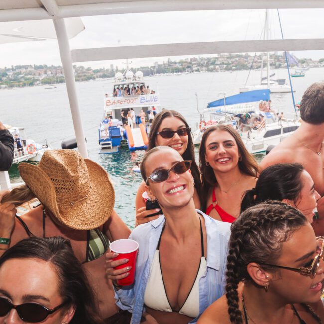 A group of young people in swimsuits smile and enjoy drinks on a crowded boat with other boats and the ocean in the background. The atmosphere is lively and festive, with sunny weather and water all around.