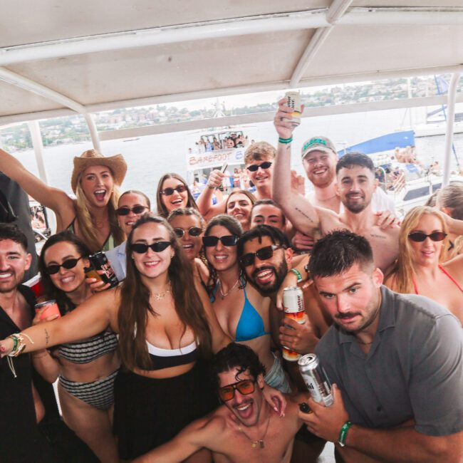 A group of young adults in swimsuits and sunglasses smile, laugh, and hold drinks while posing closely together on a boat. Water and other boats are visible in the background, suggesting a lively party atmosphere.