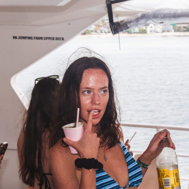 A woman in a striped bikini holding a drink and a bottle stands on a boat with others. She has wet hair, a scrunchie on her wrist, and is making a playful expression. Water and houses are visible in the background.