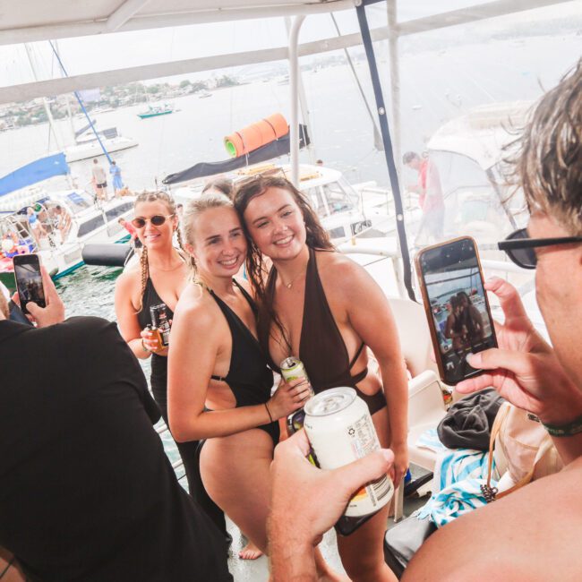 Two women in swimsuits pose and smile for photos on a boat while others take pictures. People are holding drinks, and sailboats and water are visible in the background, suggesting a lively outdoor party atmosphere.