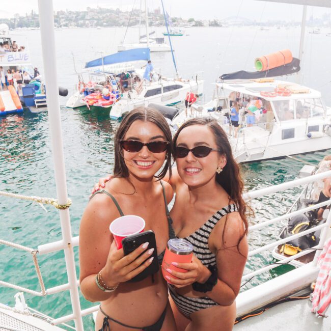 Two women in swimsuits and sunglasses smile and pose with drinks on a boat. Behind them, other boats and people are visible on the water, giving a festive, summertime atmosphere.