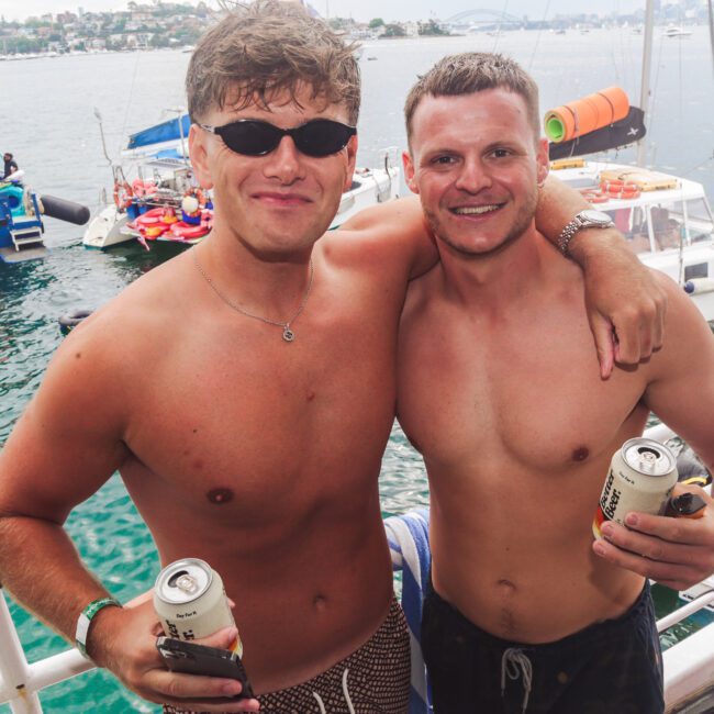 Two smiling men in swim trunks stand arm-in-arm on a boat, each holding a canned drink. Several boats and people are visible on the water behind them, with a cityscape and bridge in the distance.