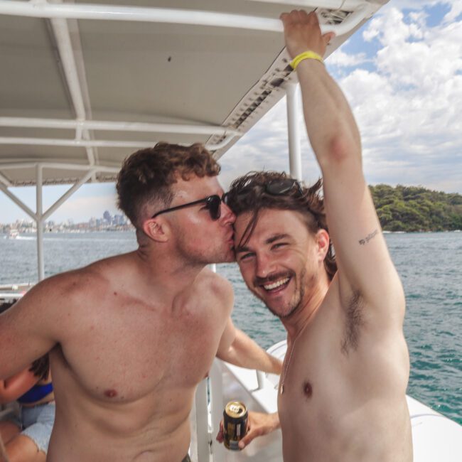 Two shirtless men smile on a boat; one kisses the other on the cheek. They're surrounded by water with trees and buildings in the background. Other people relax nearby. It’s a sunny day with scattered clouds in the sky.