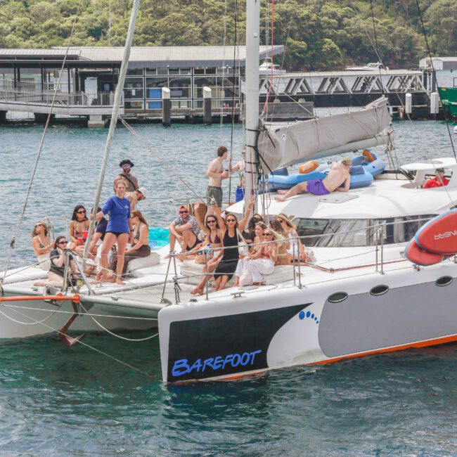 A group of people are relaxing and posing for photos on a white catamaran named "Barefoot" anchored near a dock, with trees and buildings in the background. Some are sitting, while others stand on the deck and netting.
