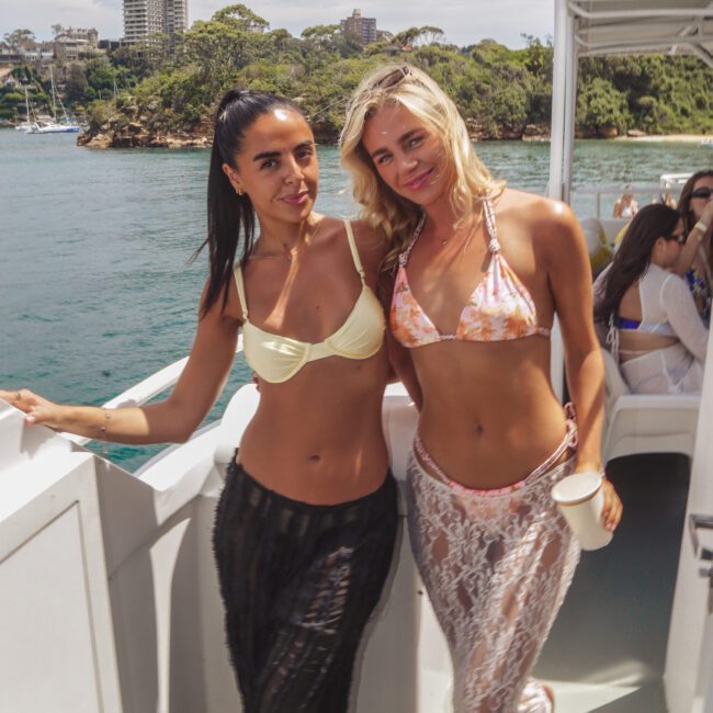 Two women in bikinis and sheer cover-ups pose together on the deck of a boat, with water, trees, and city buildings in the background under a cloudy sky. Other people are seated nearby.