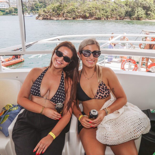 Two women in swimsuits and sunglasses sit and smile on a boat, each holding a can drink. There is water and greenery in the background, and people can be seen on another boat behind them.