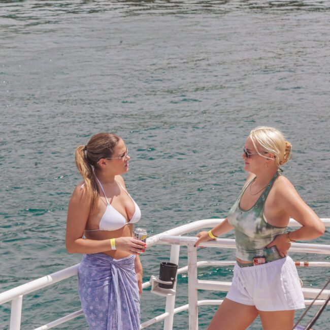 Two women stand on a boat, talking and smiling. One wears a white bikini top and wrap skirt, the other a green tank top and white shorts. The ocean is in the background. Both wear sunglasses and wristbands.