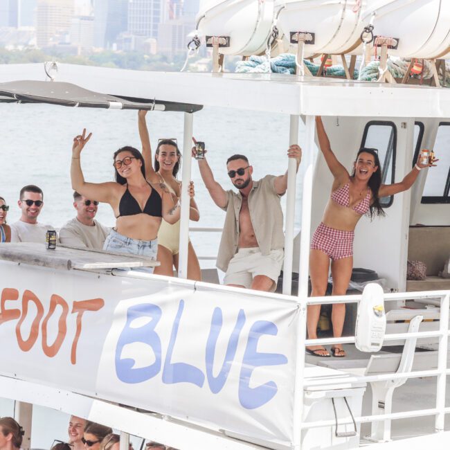 A group of smiling people in swimsuits and summer clothes enjoy drinks on the deck of a boat labeled "BAREFOOT BLUE," with water and a city skyline in the background. Some are waving and posing for the camera.