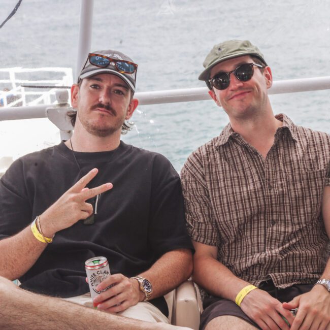 Two men sit side by side on a boat. One makes a peace sign and holds a can, both wear sunglasses, hats, and wristbands. The ocean and part of the boat are visible in the background.