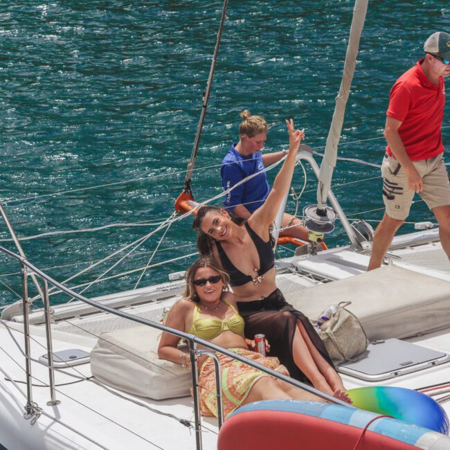 Two women lounge and smile on the deck of a sailboat, one flashing a peace sign. Three other people, two seated and one standing, enjoy the sunny day on the sparkling blue water.