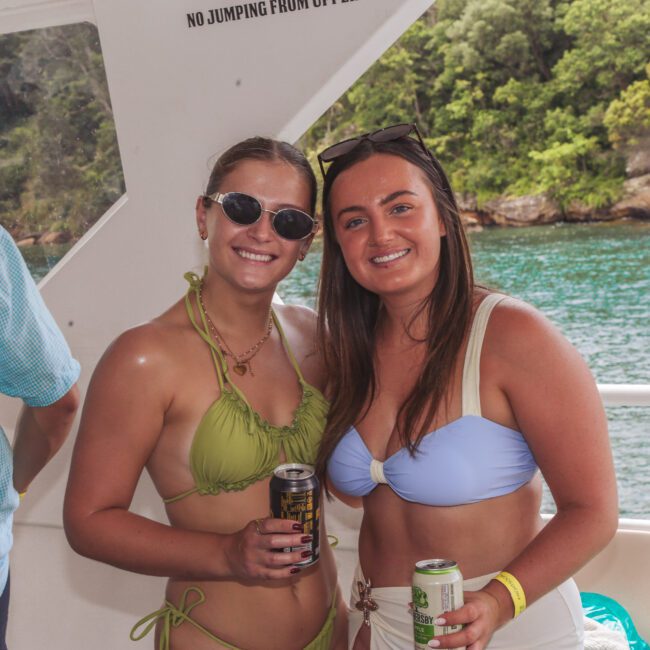 Two women in swimsuits, sunglasses, and wristbands smile and hold drinks on a boat. There is water and greenery visible through the window, and other people around them.