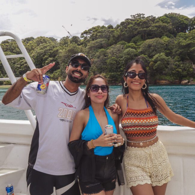 Three people smile and pose on a boat in front of a lush, green island. Two women wear summer outfits and sunglasses; the man wears a cap and sunglasses, holding a drink. The water is turquoise and the sky partly cloudy.
