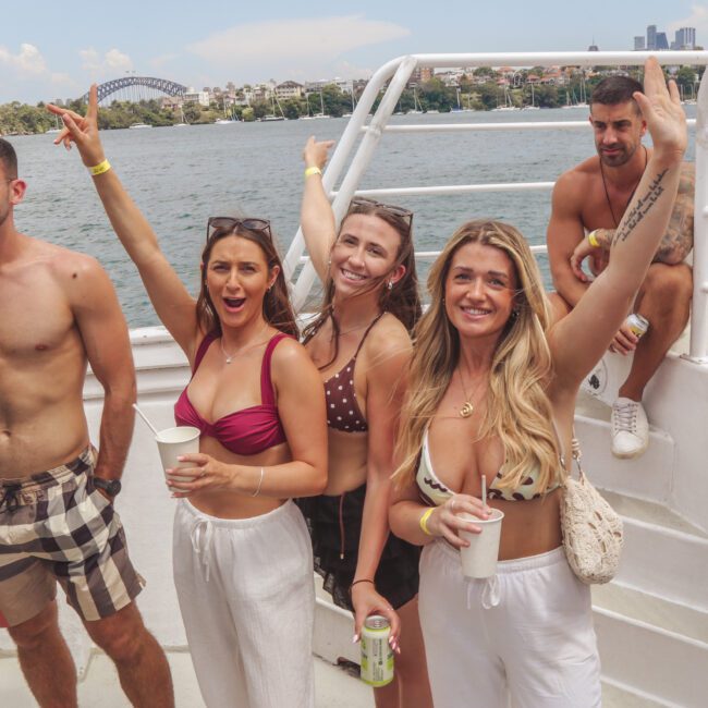 A group of five young adults in swimwear smile and pose on a boat with drinks in hand; the Sydney Harbour Bridge and city skyline are visible in the background under a sunny sky.